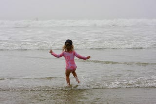 A small child has her back to the camera. She is dancing towards waves on the ocean.