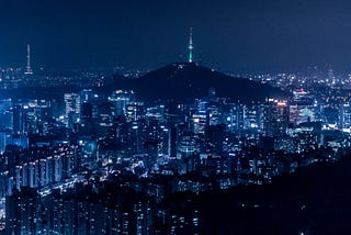 A night time photo of the city of Seoul Korea. In blue and black colours with lots of white lights.