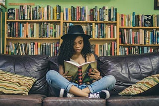 A young woman in a hat and blue jeans reading a book. She is sitting on a black, leather couch with her legs crossed. She is indoors, with a row of bookshelves behind her.