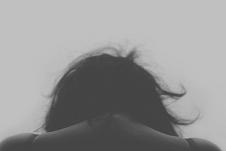A woman holds her head down. Her shoulders are up. She is sad. The photo is in greyscale.