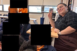 Me, the author, sitting at a desk, smiling and holding up a peace sign. Students, also in the picture, have their faces censored, but the SA logo is visible on their shirts and sweaters.
