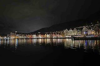 A nighttime waterfront view of Bryggen in Bergen, Norway, featuring a row of brightly lit, colorful wooden buildings with steep gabled roofs. Their reflections shimmer on the calm water in front. Behind the historic harbor district, a hillside dotted with lights from homes rises into the dark sky, adding depth to the scenic and culturally rich landscape.
