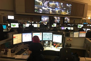 A large room filled with desks. Each desk holds several monitors. On the wall, a large screen is playing what appears to be security camera footage.