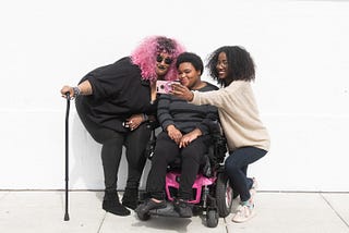 A non-binary person with a cane, a non-binary person sitting in a power wheelchair, and an invisibly disabled femme smiling and taking a cell phone selfie together.