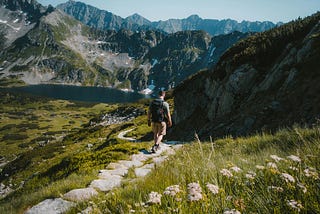 Photo by Josh Hild — a lone walker, walking off into the distance along a winding stone path in the countryside, with a range of mountains in the distance. The sun is shining, and the grass is really green and vibrant. A few wildflowers are showing in the foreground.