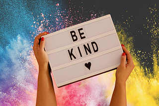 Hands with red fingernails holding a sign that says “be kind” in front of a rainbow-colored background