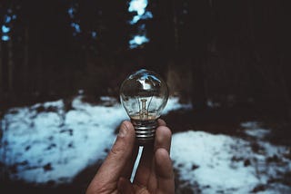 A hand holding a small lightbulb in a snowy forest.