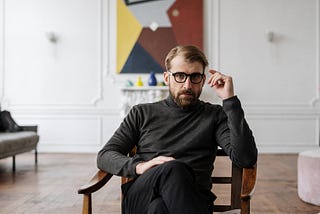 A man in glasses sitting in a chair in a room.