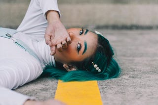 An Asian woman with black and dark green hair is lying on the street. There is a yellow street line under her head.