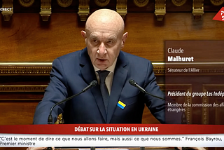 A bald man in a dark blue suit and tie is speaking at a podium in a formal government setting. He has a serious expression and is flanked by two microphones. A small pin with the colors of the Ukrainian flag is attached to his suit lapel. The background is ornate wooden paneling. The overlay text identifies him as Claude Malhuret, a senator from Allier and president of the political group Les Indépendants.