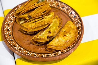 A brown ceramic plate with five crispy tacos on a yellow and white striped tablecloth.