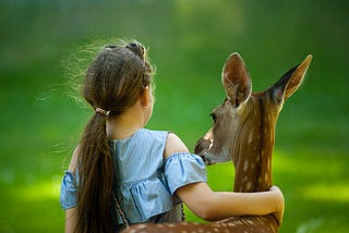 Child wearing a blue top with her arm around a young deer in the park.