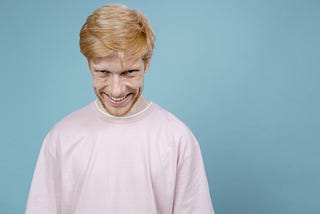 Image shows red-haired man in pink shirt against a blue background, grinning maniacally at camera.