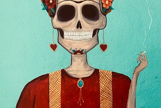 A mural on a blue wall. The mural depicts a sugar skull — a female-appearing skeleton covered in flowers and hearts. She is smoking a cigarette and grinning. Her unibrow suggests that she is Frida Khalo, or at least an homage to her.