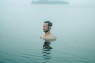 A photo of a white, Nordic-looking man standing in what appears to be a very still lake. The water comes up to just below the man’s shoulders and a dark shadowy reflection of his can be seen on the water towards the camera. The air is misty and the sky overcast and dimly lit with a haze. A blurry, dark horizontal shape that seems to be a small island of trees can be seen of in the distance on the horizon.