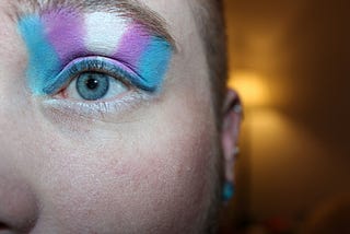 Close up photograph of half of a face of a caucasian youth, wearing studs earrings, and shorn dirty blond hair. The photo is cenbtered on their half of their face, one blue eye and eyebrow. Above the eyebrow is the trans flag in eyeshadow extending to the lower eyebrow.