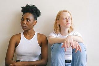 Two women seated, one White and one Black, looking vaguely unhappy.