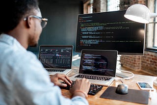 Photo of a Black person with glasses sitting in front of two laptops and a desktop computer, looking at code on the screens.