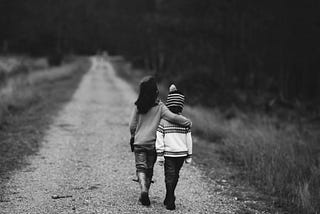 Black and white, two children walking down a road. Taller child has arm around shorter child.