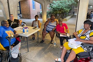 Six women sitting around a table outside an apartment building looking at the camera.