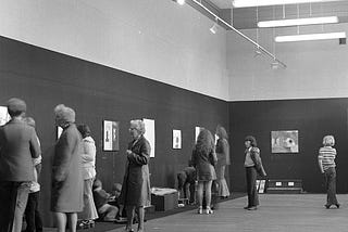A black and white photo showing a scattered crowd looking at some artworks hung on a gallery wall