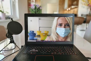 A laptop and webcam sit on a table. On the laptop screen a woman wearing a mask stares back to the viewer.