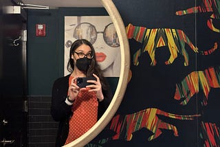 Amy holding her smartphone in front of a round mirror, with a print of a woman in sunglasses with red lipstick and colorful tiger wallpaper.
