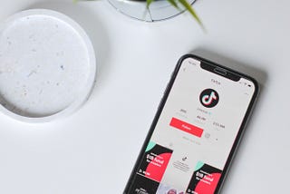 Smartphone showing the TikTok profile page. Set on a white table next to a white coaster and small potted plant.