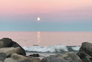 A sunset over crashing waves on a rocky shore. The sky is blue and pink. The sun is a yellow ball.