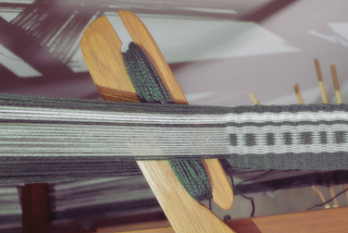 An antique weaving loom with moss green and white yarn woven into an intricate pattern.