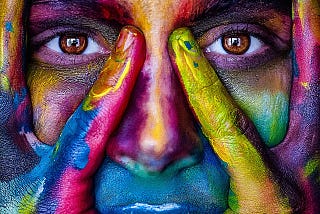 Close-up of a face with hands framing the eyes, covered in vivid, multicolored paint. The eyes are prominently visible, surrounded by bold hues of red, yellow, green, blue, and purple.