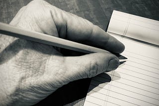 Image of a left hand holding a pencil over a pad of paper.