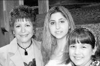 A black-and-white photograph of a mother standing beside her two daughters. All three are smiling.