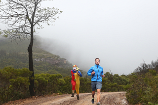 This image shows two people running on a dirt path surrounded by natural vegetation. The person in the foreground is dressed in a blue jacket and running shorts, clearly engaged in a run, looking forward with focus and determination. The other person is in a colorful clown costume, complete with a wig, face paint, and nose, and is holding what appears to be a flower. They are running slightly behind the first person and looking at the camera, adding a humorous and unexpected element to the scene