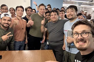 a group of smiling mostly-male Twitter employees gathered around their new boss Elon Musk at a late-night code review. Musk and others are doing a thumbs-up.