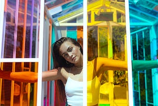 A woman with long dark hair in a white tank top casually looks through a gap in a house of colored panels