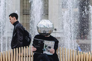 Person with a shiny disco ball as a head sitting on a bench reading an ‘ideas’ magazine while someone who looks aloof walks past in the background.