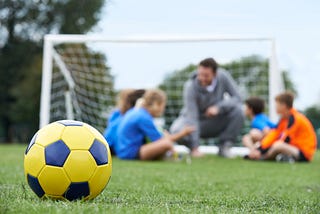 A football on a pitch with a coach and player out of focus in the background