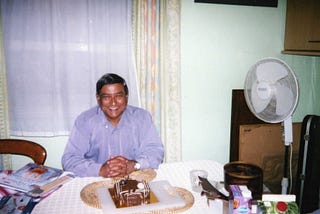 The photo shows the author’s father on his birthday. He’s a man with brown skin wearing a lilac shirt and is sat down at a table smiling in front of a chocolate birthday cake.