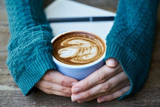 Photo of a pair of female hands nursing a cup of coffee on a rustic wooden table. The sleeves of her green jumper are pulled down level with her thumbs, as if she is cold. Or anxious.