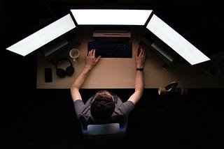 Man sitting in dark room in front of three lit up monitors. The view is from straight above him.