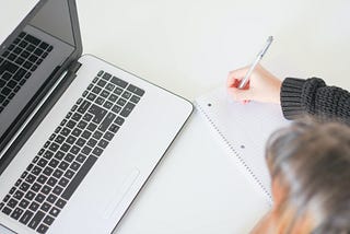 Woman taking notes on a notebook while looking at her laptop computer.