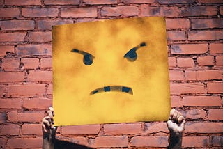 Two hands hold up a square sign with an angry face on it in front of a painted red brick wall.