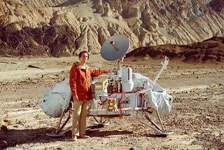 Carl Sagan stands beside a lunar lander craft in an orange jacket and beige slacks.