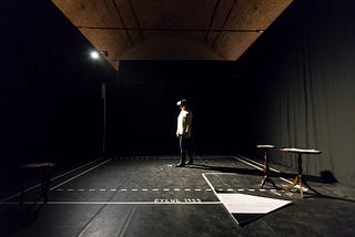 A single woman stands in the middle of a room sectioned off by floor-to-ceiling black drapes. The project details are printed on the ground, next to two tables. The woman is wearing a VR headset, a light-colored top, jeans, and black boots. There is a single spotlight illuminating the space.