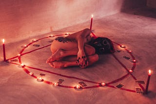 A naked woman, lying on her side, curled in a fetal position, in the centre of an occult symbol ringed with candles and tarot cards.