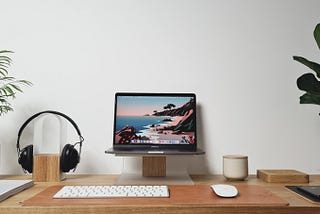 Wooden desk with a laptop perched on a stand, headphones, white wireless keyboard, and a white wireless mouse