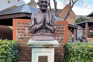 Statue of Mahatma Gandhi with his hands in prayer pose. Half a brick wall behind it and some hedges to either side. Plague reads: ‘May noble thoughts come to us from all directions” Mahatma Gandhi 1869–1948. Unveiled by Rt Hon The Lord Puttnam CBE. On the 15th October 2004.