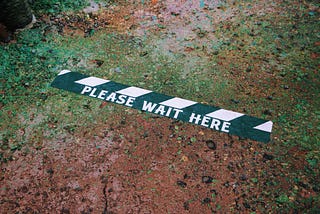 Black and white striped tape on dirt reads “Please Wait Here”