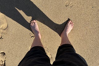 Mark’s bare feet on sand, casting a shadow to the left